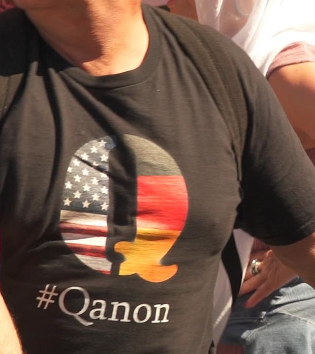 "QAnon - Wie gefährlich kann eine Verschwörungstheorie werden": Oberkörper eines Mannes der ein schwarzes T-Shirt mit geteiltem Q-Symbol, der US-Flagge und Deutschland-Flagge, und Schriftzug trägt.