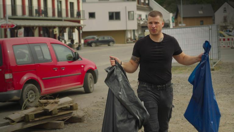 Bürgermeister David Fuhrmann räumt am Dreckwegtag an der Ahr Müll weg. Er trägt ein schwarzes T-Shirt und hält zwei Müllsäcke in der Hand.