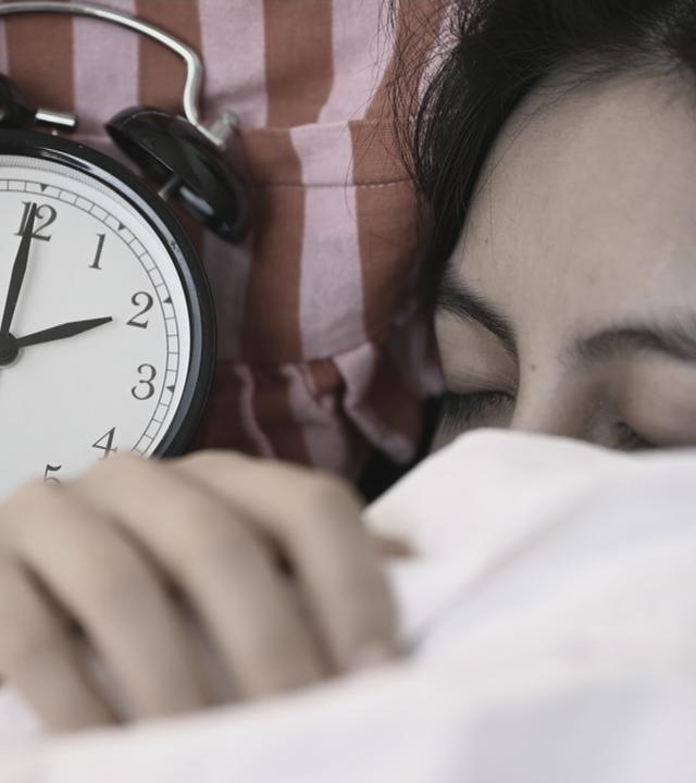 Eine Frau zieht sich die Decke über den Kopf. Neben ihr ist eine Uhr.