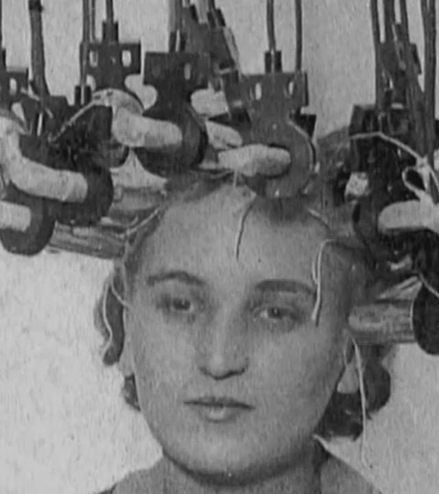 Schwarz-Weiß-Aufnahme einer Frau mit Wicklern und Haube beim Friseur