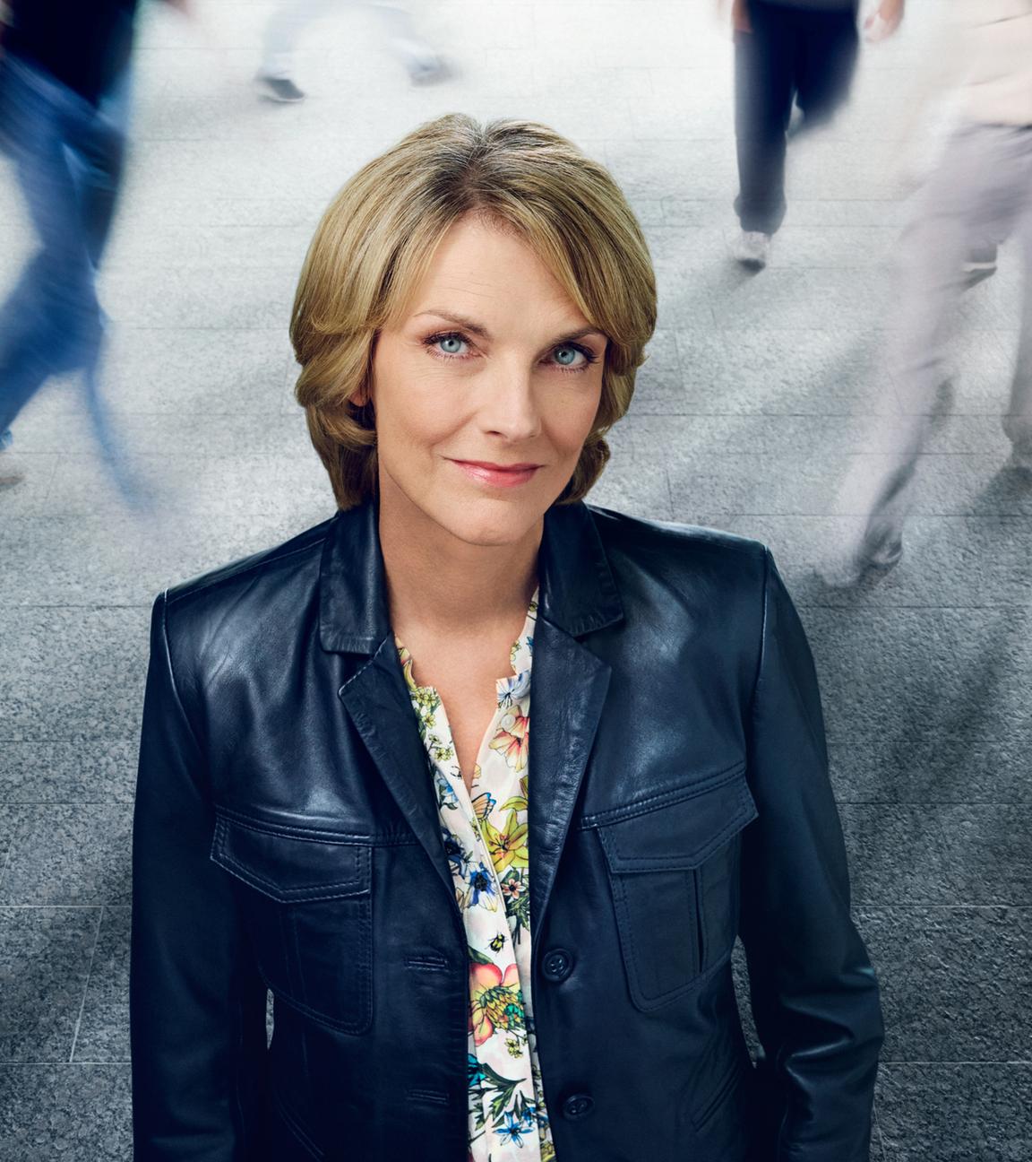 Marietta Slomka aus leicht erhöhter Position fotografiert, schaut leicht nach oben in die Kamera. Sie steht vor einer Hintergrundprojektion mit Betonfliesen und Menschen, die in Bewegungsunschärfe gehen.