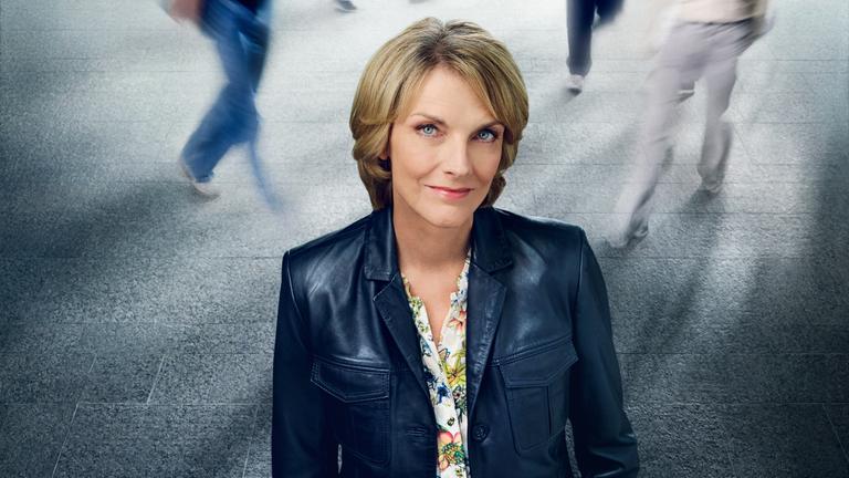 Marietta Slomka aus leicht erhöhter Position fotografiert, schaut leicht nach oben in die Kamera. Sie steht vor einer Hintergrundprojektion mit Betonfliesen und Menschen, die in Bewegungsunschärfe gehen.