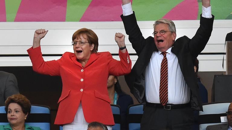 Das Bild zeigt Angela Merkel und den damaligen Bundespräsidenten Joachim Gauck jubelnd bei einem Fußballspiel.