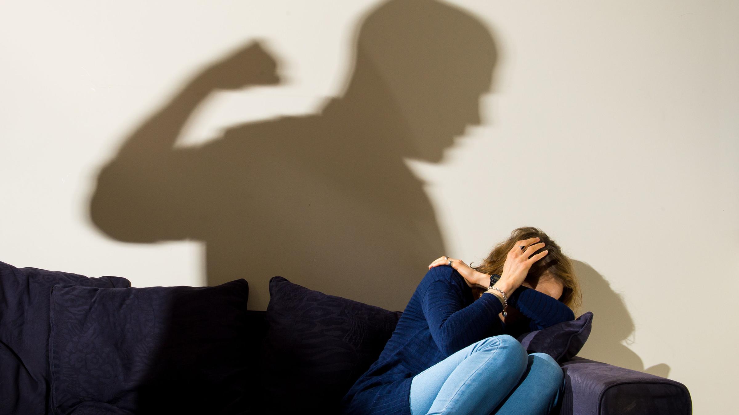 London 02.04.2021, Eine Frau hält schützend die Arme vor da sGesicht während sie auf dem Sofa sitzt und der Schatten eines Mannes mit geballter Faust zu sehen ist.