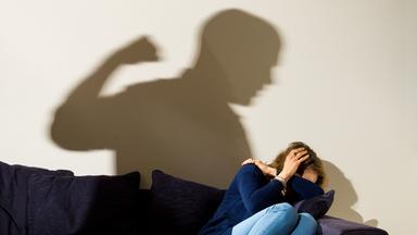 London 02.04.2021, Eine Frau hält schützend die Arme vor da sGesicht während sie auf dem Sofa sitzt und der Schatten eines Mannes mit geballter Faust zu sehen ist.
