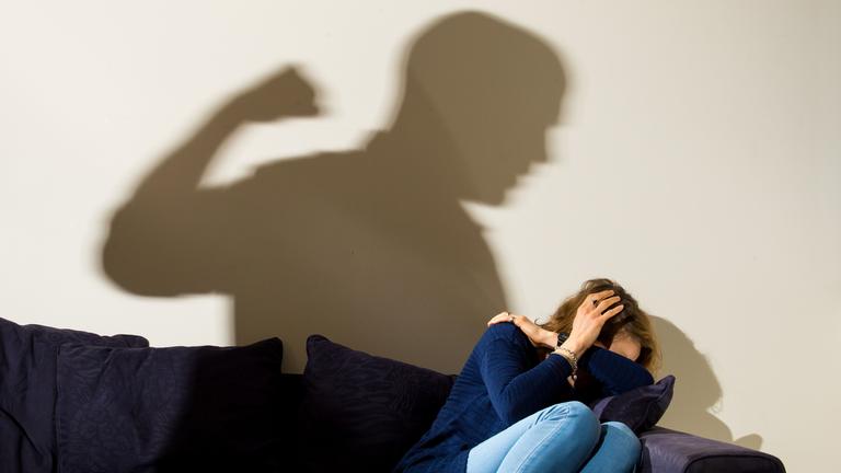 London 02.04.2021, Eine Frau hält schützend die Arme vor da sGesicht während sie auf dem Sofa sitzt und der Schatten eines Mannes mit geballter Faust zu sehen ist.