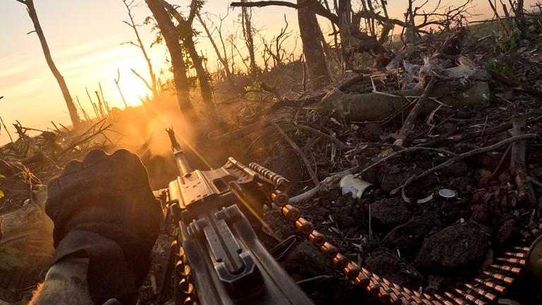 Ein ukrainischer Soldat, dessen Hand zu sehen ist, feuert mit einem Maschinengewehr auf russische Stellungen inmitten von kahlen Bäumen.