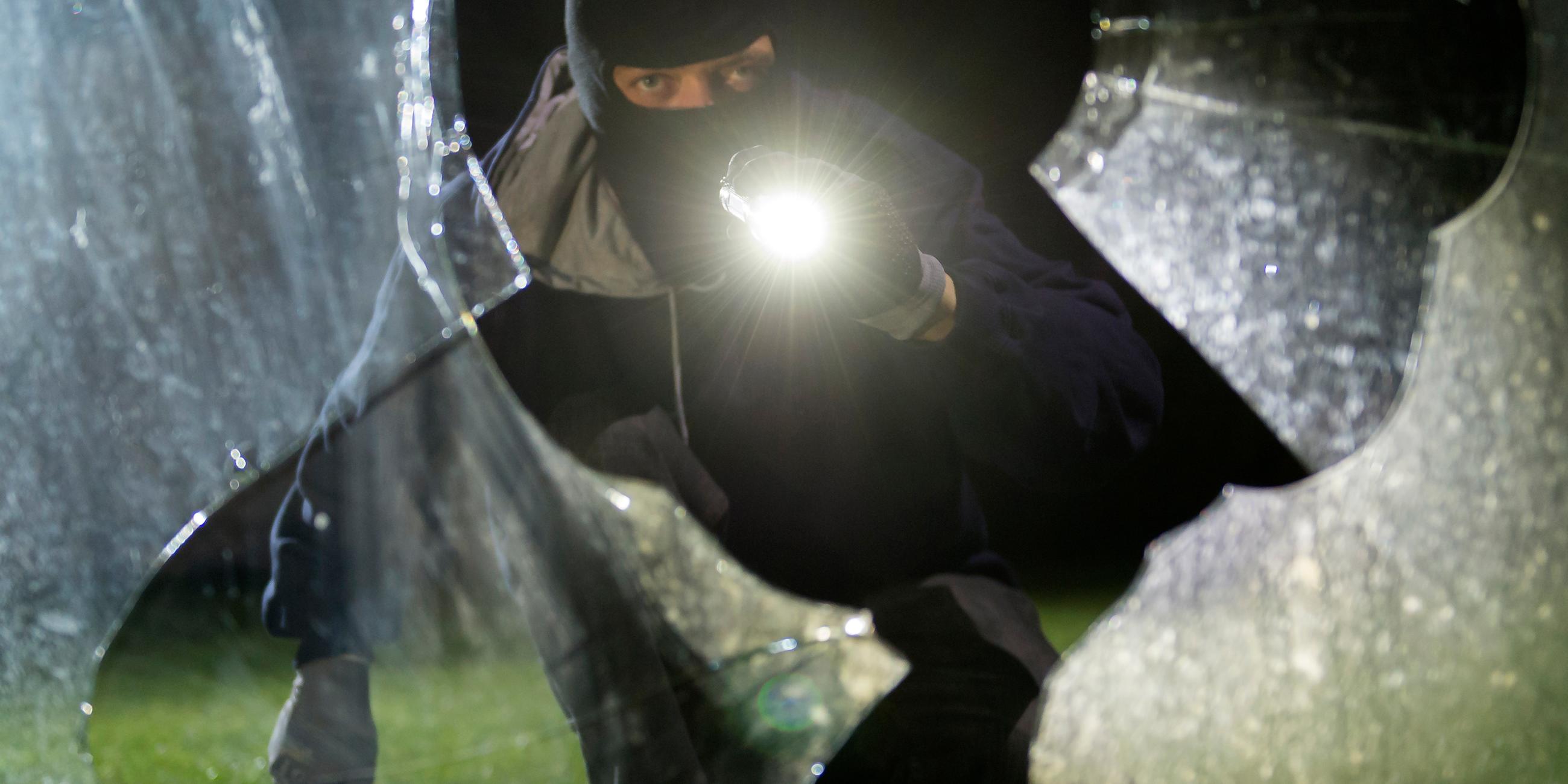 Einbrecher mit Maske und Taschenlampe leutet durch zebrochene Glasscheibe
