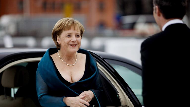Angela Merkel ist in einem Kleid mit Perlenkette und Handtasche zu sehen. 