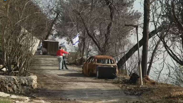 Es ist der zerstörte Kibbuz Menara zu erkennen, mit verbrannten Autos und Israel-Flaggen im Hintergrund.
