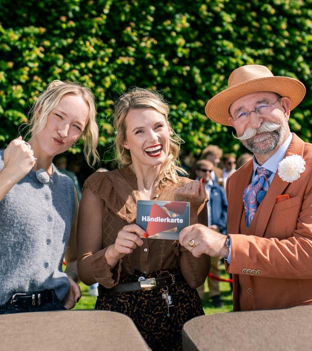 "Bares für Rares XXL": Leonie Bartsch, Linn Schütze, und Horst Lichter stehen hinter dem Expertisenpult und lächeln in die Kamera. Horst Lichter überreicht Linn Schütze die Händlerkarte. Im Hintergrund steht Publikum.