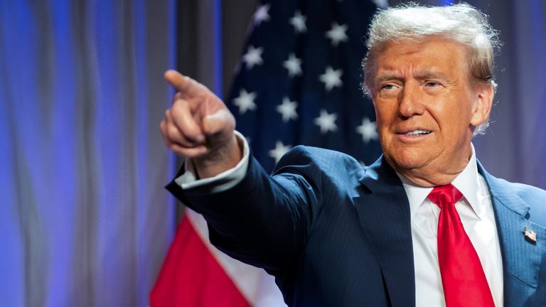 "Donald Trump - König im Weißen Haus?": Porträtaufnahme: Donald Trump steht im Anzug vor einer amerikanischen Flagge. Er zeigt mit dem rechten Arm und ausgestrecktem Zeigefinger auf etwas. Sein Blick folgt seinem Arm.