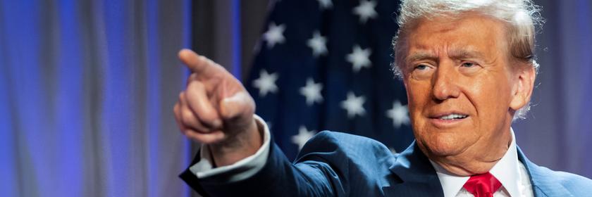 "Donald Trump - König im Weißen Haus?": Porträtaufnahme: Donald Trump steht im Anzug vor einer amerikanischen Flagge. Er zeigt mit dem rechten Arm und ausgestrecktem Zeigefinger auf etwas. Sein Blick folgt seinem Arm.