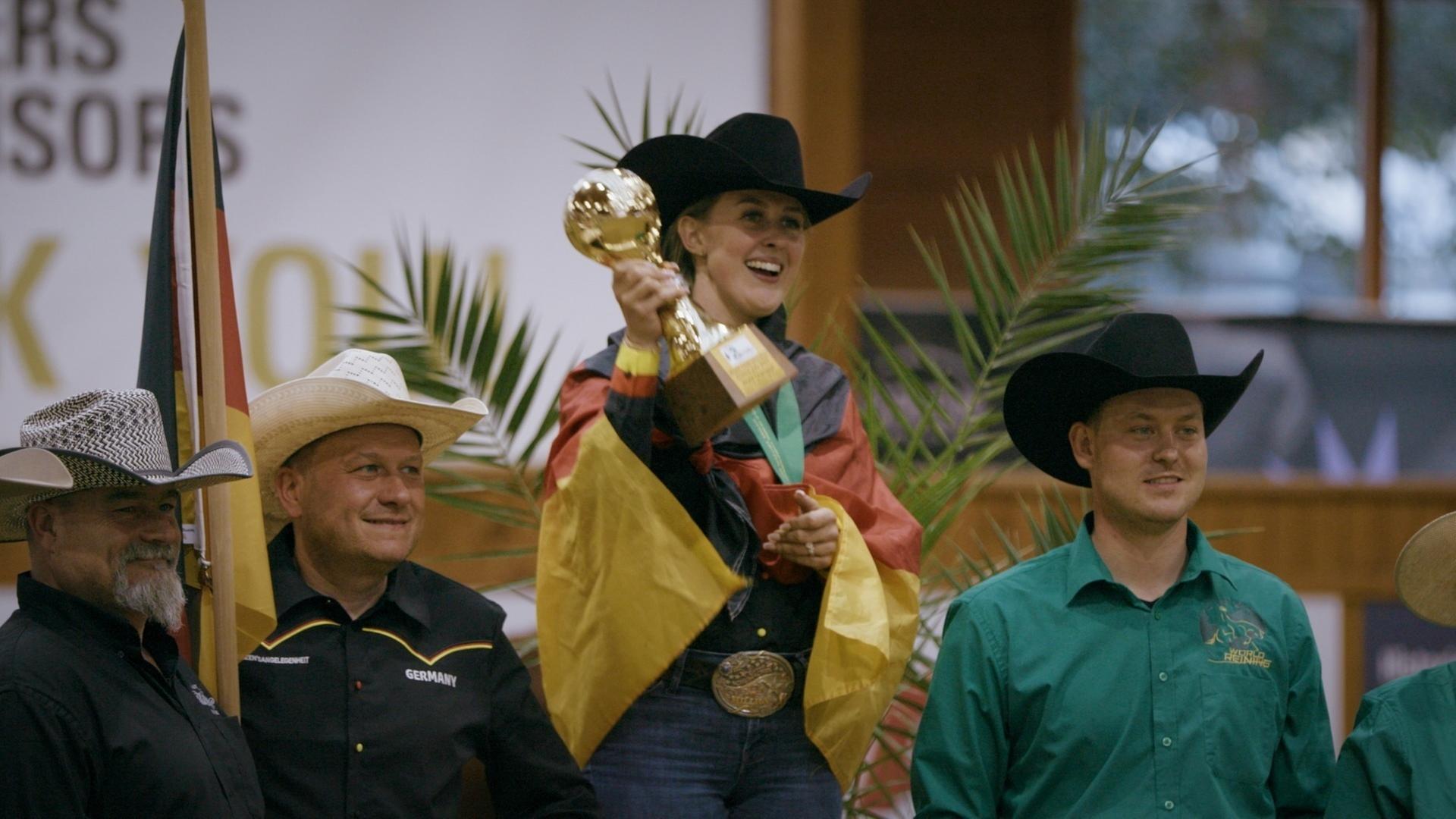 "Pferdestärke - Die Welt der Gina Schumacher": Gina Schumacher steht bei den World Reining Championships 2025 in Givrins in der Schweiz erhöht neben drei Männern und hält einen Pokal in der Hand. Sie hat eine deutsche Fahne um ihre Schultern gelegt.