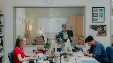 "Nine to Five - Das Amt - Ferienanlage Döberitz": Melissa (Anna Herrmann) und Orkan (Yasin El Harrouk) sitzen sich an ihren jeweiligen Schreibtischen gegenüber und blicken auf Bildschirme. Im Hintergrund schaut Ronja (Friederike Kempter) durch ihr Bürofenster auf die beiden.