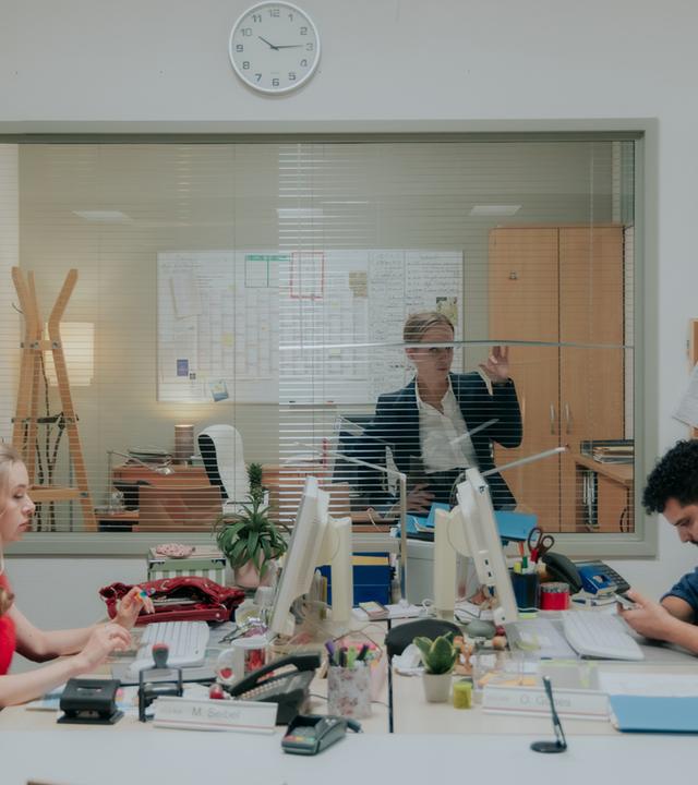"Nine to Five - Das Amt - Ferienanlage Döberitz": Melissa (Anna Herrmann) und Orkan (Yasin El Harrouk) sitzen sich an ihren jeweiligen Schreibtischen gegenüber und blicken auf Bildschirme. Im Hintergrund schaut Ronja (Friederike Kempter) durch ihr Bürofenster auf die beiden.