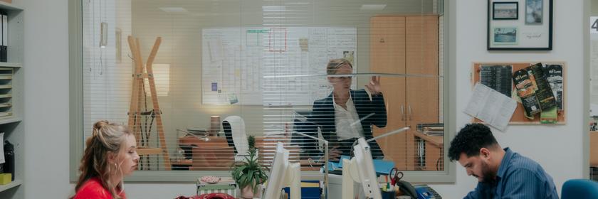 "Nine to Five - Das Amt - Ferienanlage Döberitz": Melissa (Anna Herrmann) und Orkan (Yasin El Harrouk) sitzen sich an ihren jeweiligen Schreibtischen gegenüber und blicken auf Bildschirme. Im Hintergrund schaut Ronja (Friederike Kempter) durch ihr Bürofenster auf die beiden.