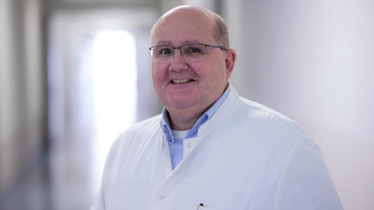 Prof. Dr. Bernhard Schieffer steht in einem Medizinerkittel in einem Krankenhausflur. Er trägt eine Brille und hat eine Halbglatze 