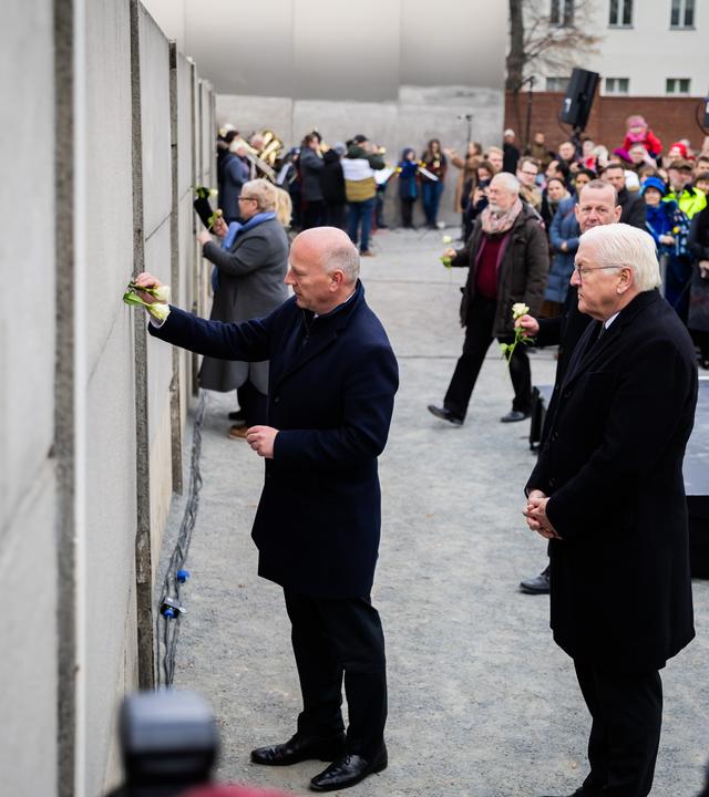 Kai Wegner (CDU, l), Regierender Bürgermeister von Berlin, steckt neben Bundespräsident Frank-Walter Steinmeier und Lisa Paus (Bündnis 90/Die Grünen), Bundesministerin für Familie, Senioren, Frauen und Jugend, bei einer zentralen Gedenkveranstaltung anlässlich von 35 Jahre friedliche Revolution und Mauerfall in der Gedenkstätte Berliner Mauer eine Rose in die Hinterlandmauer. 
