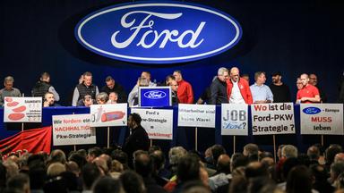 Mitarbeiter, Gewerkschaftler stehen auf einer Bühne mit Schildern, dahinter ein Ford Logo