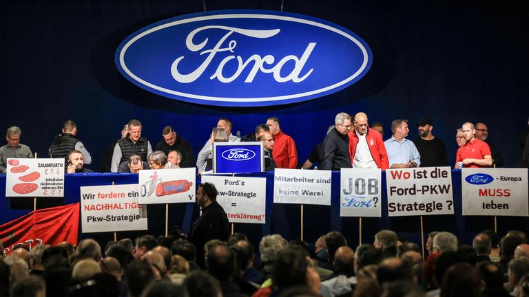 Mitarbeiter, Gewerkschaftler stehen auf einer Bühne mit Schildern, dahinter ein Ford Logo