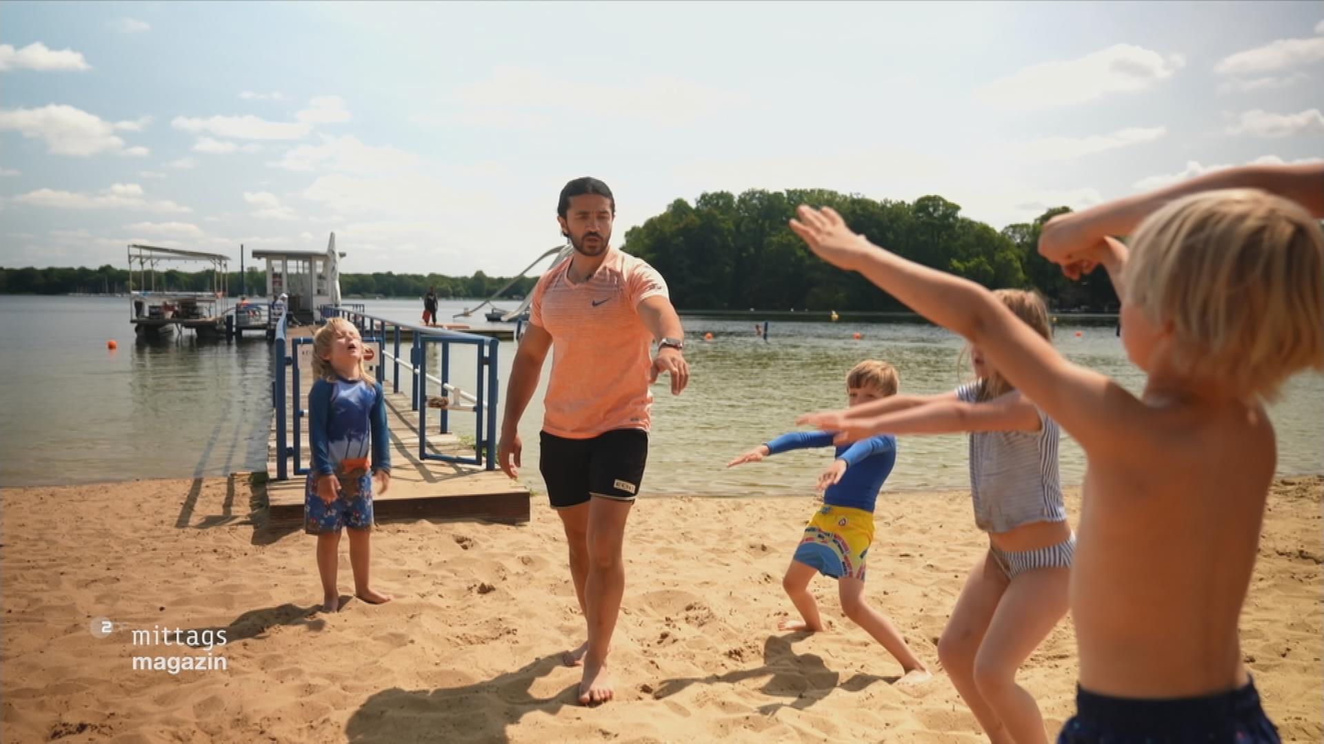 Schwimmlerher bringt Kindern am Steg vorm Ufer Bewegungen bei
