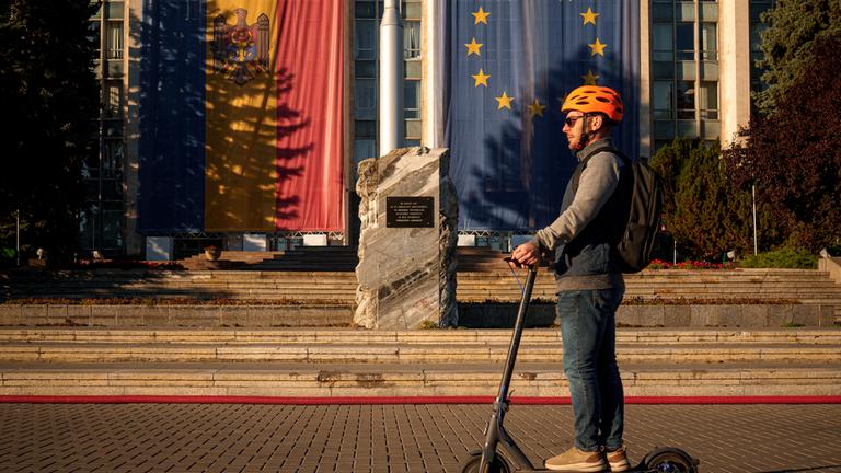 Mann auf einem Elektroroller vor Regierungsgebäude mit Moldau- und Europaflagge