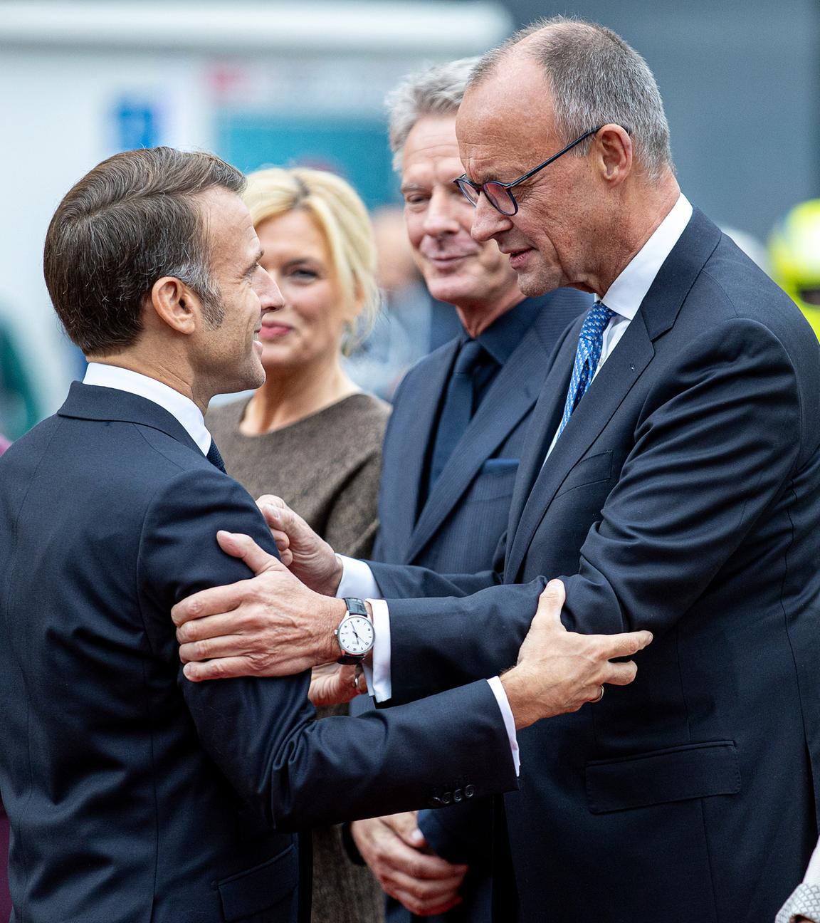 Frankreichs Präsident Emmanuel Macron (M.l) begrüßt Bundeskanzler Friedrich Merz (CDU) bei seiner Ankunft vor der saarländischen Staatskanzlei während der zentralen Feierlichkeiten dem Tag der Deutschen Einheit, Brigitte Macron, Anke Rehlinger (SPD), saarländische Ministerpräsidentin und Bundesratspräsidentin, Bundestagspräsidentin Julia Klöckner (CDU) und ihr Partner, der Fernsehmoderator Jörg Pilawa, sowie Charlotte Merz, Frau des Bundeskanzlers, sehen zu. Am 3. Oktober 2025 begeht Deutschland den 35. Jahrestag der deutschen Wiedervereinigung