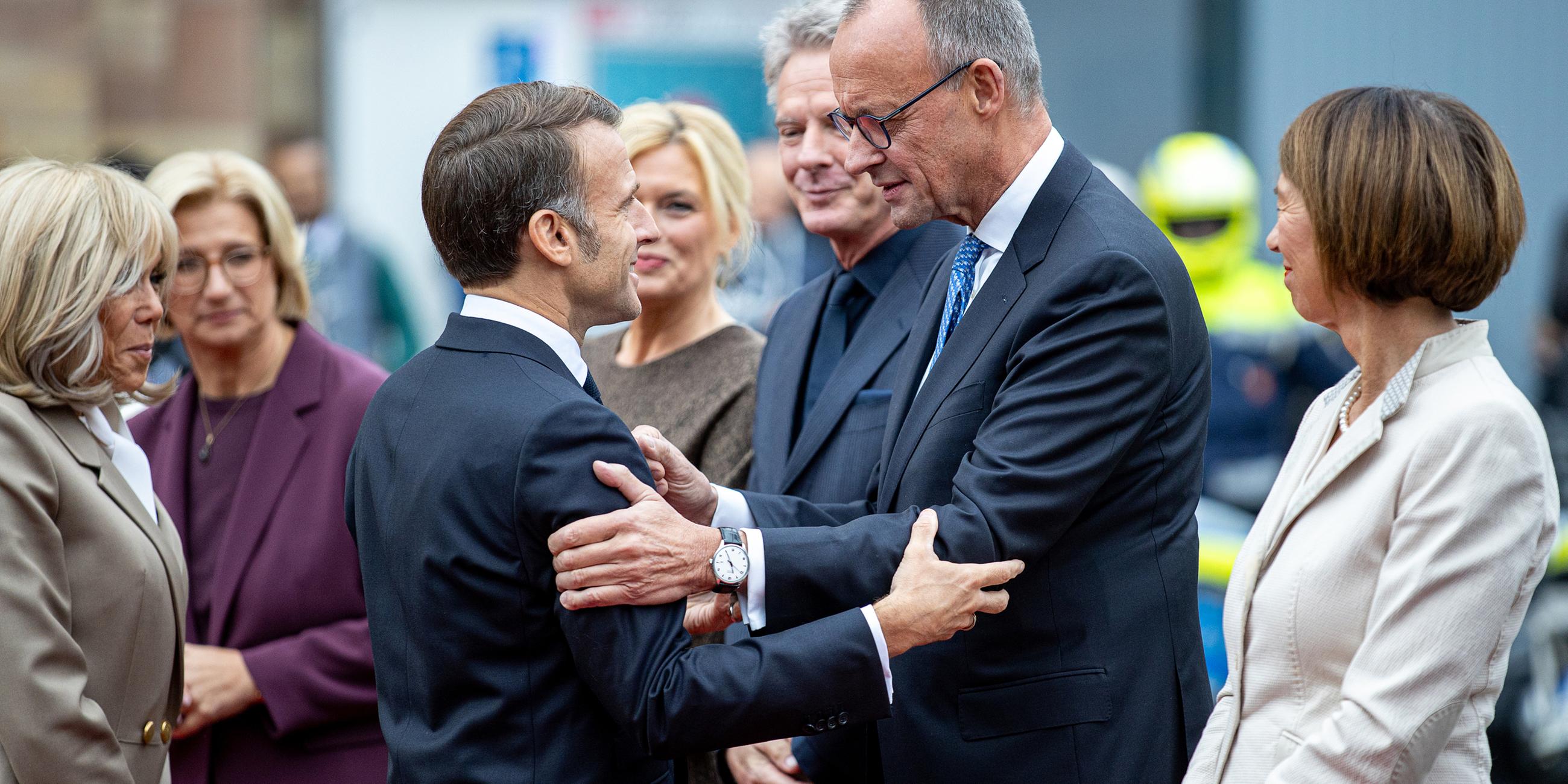 Frankreichs Präsident Emmanuel Macron (M.l) begrüßt Bundeskanzler Friedrich Merz (CDU) bei seiner Ankunft vor der saarländischen Staatskanzlei während der zentralen Feierlichkeiten dem Tag der Deutschen Einheit, Brigitte Macron, Anke Rehlinger (SPD), saarländische Ministerpräsidentin und Bundesratspräsidentin, Bundestagspräsidentin Julia Klöckner (CDU) und ihr Partner, der Fernsehmoderator Jörg Pilawa, sowie Charlotte Merz, Frau des Bundeskanzlers, sehen zu. Am 3. Oktober 2025 begeht Deutschland den 35. Jahrestag der deutschen Wiedervereinigung