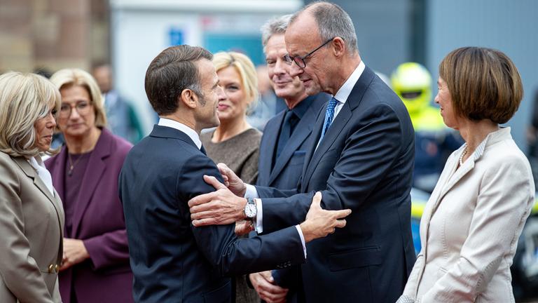 Frankreichs Präsident Emmanuel Macron (M.l) begrüßt Bundeskanzler Friedrich Merz (CDU) bei seiner Ankunft vor der saarländischen Staatskanzlei während der zentralen Feierlichkeiten dem Tag der Deutschen Einheit, Brigitte Macron, Anke Rehlinger (SPD), saarländische Ministerpräsidentin und Bundesratspräsidentin, Bundestagspräsidentin Julia Klöckner (CDU) und ihr Partner, der Fernsehmoderator Jörg Pilawa, sowie Charlotte Merz, Frau des Bundeskanzlers, sehen zu. Am 3. Oktober 2025 begeht Deutschland den 35. Jahrestag der deutschen Wiedervereinigung
