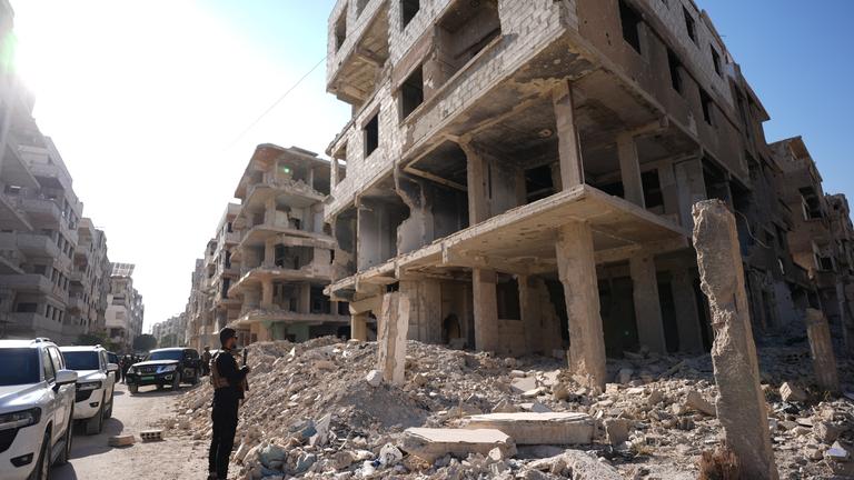 Sicherheitskräfte aus Syrien und aus Deutschland sichern den Besuch von Außenminister Wadephul bei einem humanitären Projekt in einer Straße mit während des Bürgerkriegs zerbombten Häusern ab