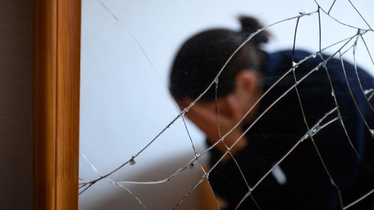 Man sieht eine Frau vor einem gesplitterten Spiegel, wie sie ihr Gesicht in den Händen hält.