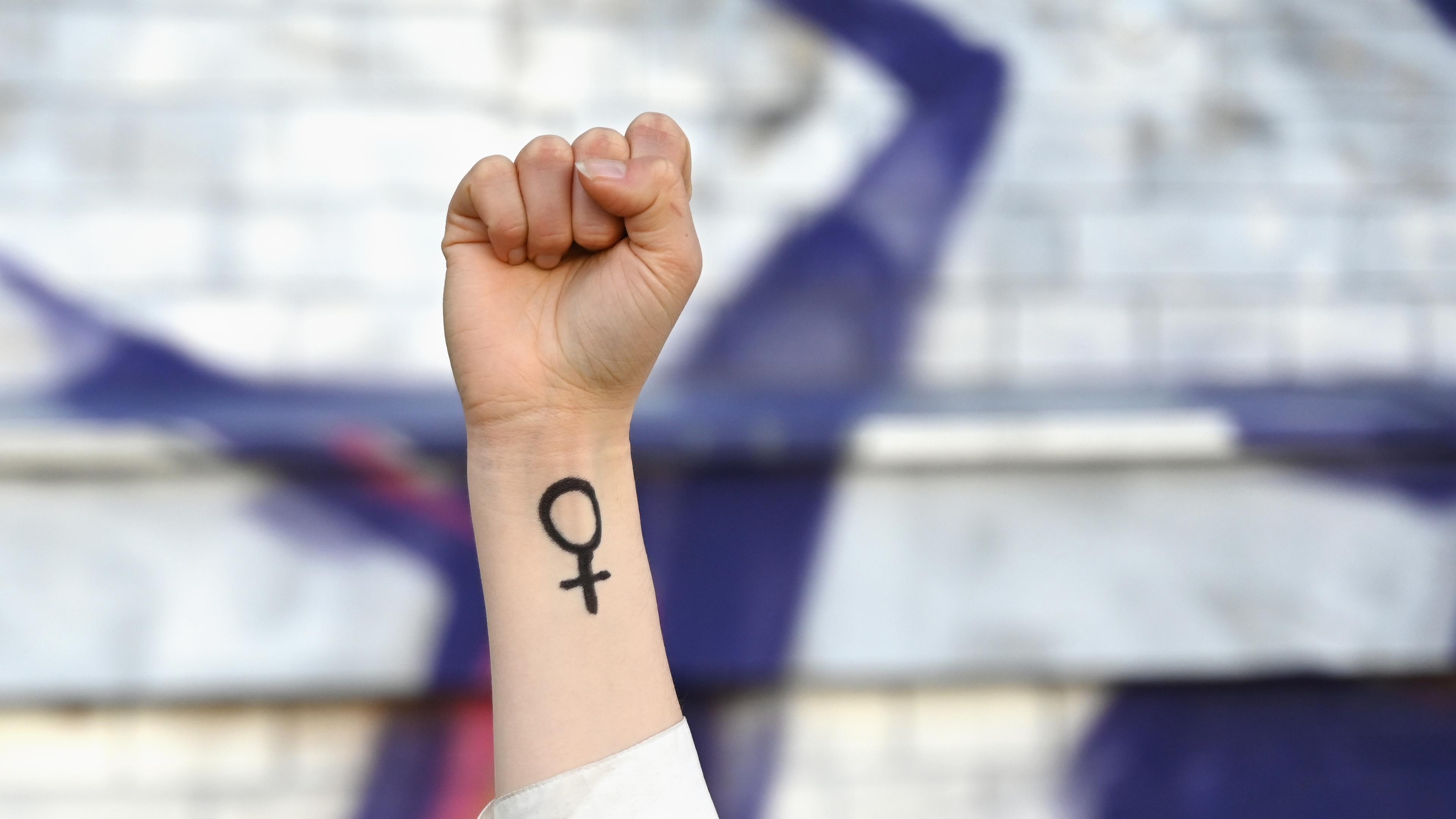 Eine Frau hebt ihre Faust in die Höhe. Auf ihrem Arm ist das Venussymbol zu sehen, das für das weibliche Geschlecht und Frauenrechte steht. 