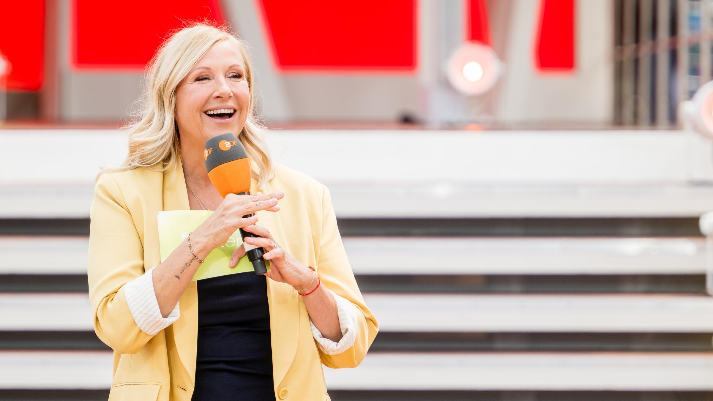 Andrea Kiewel steht mit gelber Jacke und Mikrofon in der Hand vor der Fernsehgarten-Showtreppe.