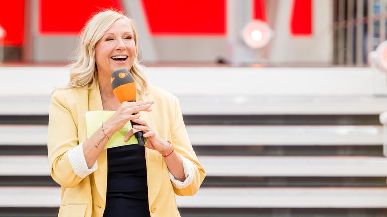 Andrea Kiewel steht mit gelber Jacke und Mikrofon in der Hand vor der Fernsehgarten-Showtreppe.