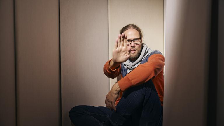 René (39) sitzt in einer Ecke auf dem Boden. Er hält seine rechte Hand abwehrend und schützend vor sein Gesicht.