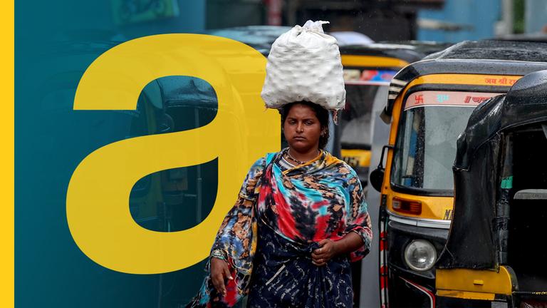 Man erkennt eine indische Frau, die auf dem Kopf Waren trägt zwischen Tuktuks.