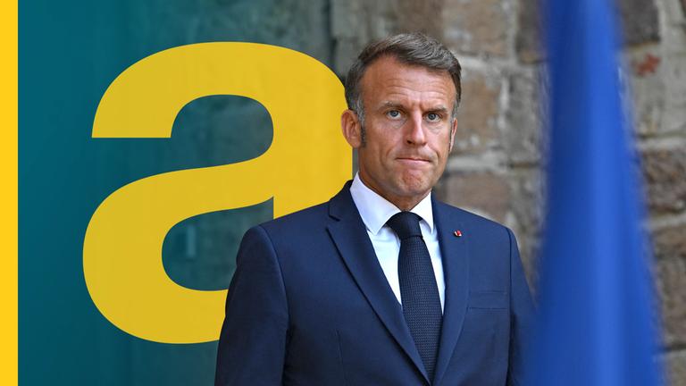 Emmanuel Macro vor einer Steinwand, hat die Lippen zusammengepresst - im Vordergrund unscharf eine blaue Fahne.