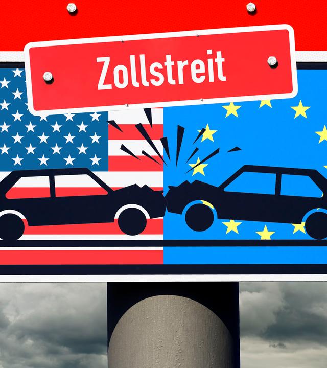 Zusammenstoßende Autos vor den Fahnen von den USA und Europa auf einem Verkehrsschild mit Aufschrift Zollstreit