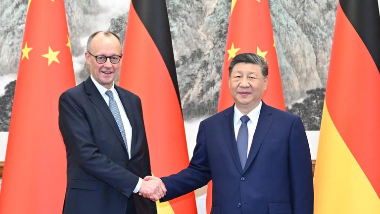 Merz und Xi Jinping reichen sich vor ihren Landesflaggen die Hand