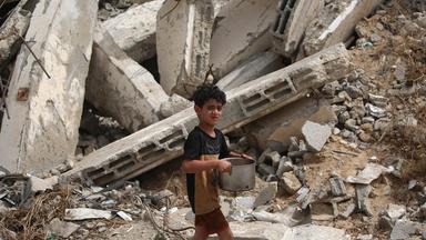 Ein palästinensisches Flüchtlingskind trägt im Flüchtlingslager Bureij im zentralen Gazastreifen einen Topf mit Essen, das von einer Suppenküche ausgegeben wurde, vorbei an Gebäuden, die vom israelischen Militär zerstört wurden.