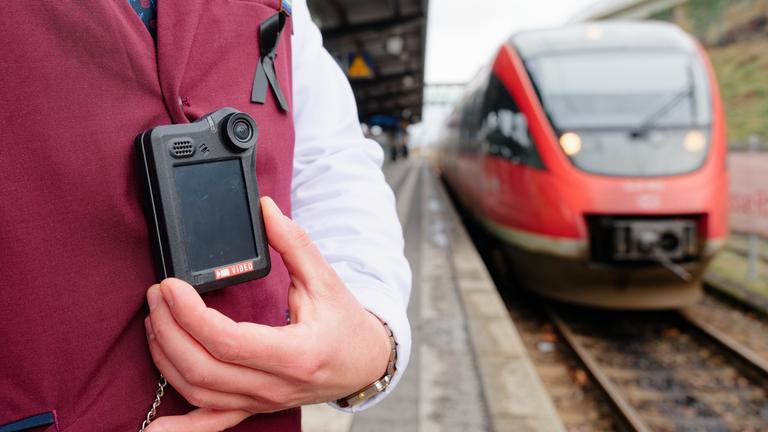 13.02.2026, Rheinland-Pfalz, Neustadt an der Weinstraße: Ein Kundenbetreuer steht bei einem Pressetermin der Deutsche Bahn (DB) auf einem Bahnsteig neben einem Regionalzug und trägt eine Bodycam an der Dienstkleidung. 