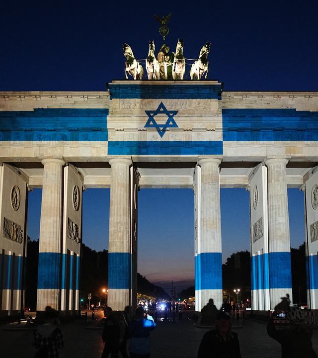 07.10.2026, Berlin: Teilnehmer stehen vor dem mit der Fahne Israels angestrahlten Brandenburger Tor und gedenken bei der Veranstaltung zum Jahrestag des Terrorangriffs der Hamas auf Israel am 7. Oktober 2023 den Opfern.