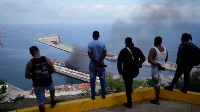 Männer beobachten, wie Rauch von einem Dock aufsteigt, nachdem im Hafen von La Guaira, Venezuela, am Samstag, 3. Januar 2026, Explosionen zu hören waren.