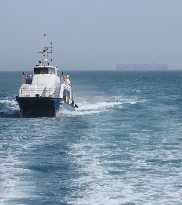 02.05.2026, Iran, Bandar Abbas: Ein Patrouillenboot fährt durch das Wasser, während Frachtschiffe in der Straße von Hormuz vor Bandar Abbas, Iran, vor Anker liegen.