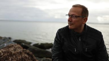 Alexander sitzt auf Steinen am Ufer der Ostsee und schaut in die Ferne. Der Himmel ist bewölkt.