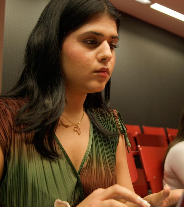 Samira, aus der Doku "37°Leben - Deutschland, wann bin ich genug?", sitzt vor einem Laptop in einem Hörsaal. Sie blickt konzentriert auf den Bildschirm. Im Hintergrund sitzen weitere Studierende auf roten Hörsaalsitzen. Der Raum ist dunkel gehalten und wird von warmem Licht beleuchtet.