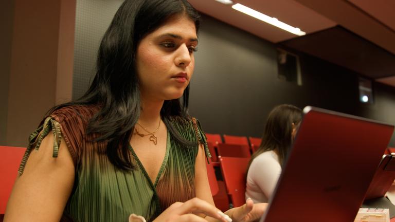 Samira, aus der Doku "37°Leben - Deutschland, wann bin ich genug?", sitzt vor einem Laptop in einem Hörsaal. Sie blickt konzentriert auf den Bildschirm. Im Hintergrund sitzen weitere Studierende auf roten Hörsaalsitzen. Der Raum ist dunkel gehalten und wird von warmem Licht beleuchtet.