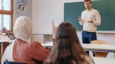 Ein Mann steht in einem Klassenzimmer und spricht zu Schülern, die mit dem Rücken zur Kamera sitzen.