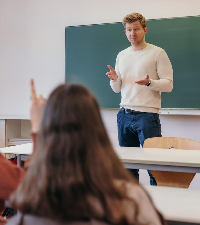 Ein Mann steht in einem Klassenzimmer und spricht zu Schülern, die mit dem Rücken zur Kamera sitzen.